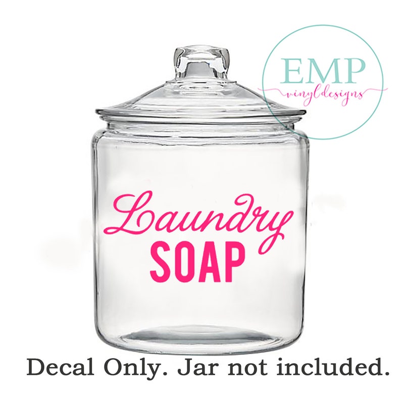 Puede incluir: Un tarro de cristal transparente con tapa, con las palabras "Laundry SOAP" en escritura rosa brillante. La calcoman&iacute;a est&aacute; dise&ntilde;ada para la organizaci&oacute;n del lavadero. La imagen tambi&eacute;n incluye un logotipo en un c&iacute;rculo que dice "EMP vinyl designs".