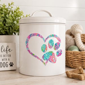 May include: A white cylindrical metal canister with a lid and handle, featuring a colorful heart-shaped graphic containing a paw print. The design is filled with a vibrant, multi-colored abstract pattern in shades of pink, blue, and green.