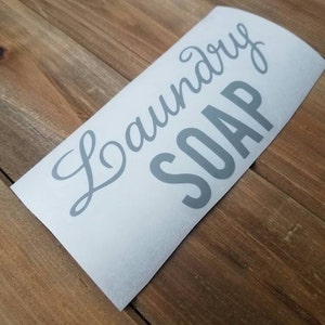 Puede incluir: Una calcoman&iacute;a de vinilo blanca con las palabras "Laundry Soap" en letras grises. La calcoman&iacute;a est&aacute; en &aacute;ngulo sobre una superficie de madera.