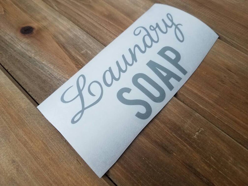 Puede incluir: Una calcoman&iacute;a de vinilo blanca con las palabras "Laundry Soap" en letras grises. La calcoman&iacute;a est&aacute; en &aacute;ngulo sobre una superficie de madera.