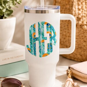May include: A white insulated tumbler with a handle and a clear lid featuring a straw. The front of the tumbler displays a circular monogram with the letters HER, filled with a vibrant peacock feather pattern in shades of teal, blue, and orange.