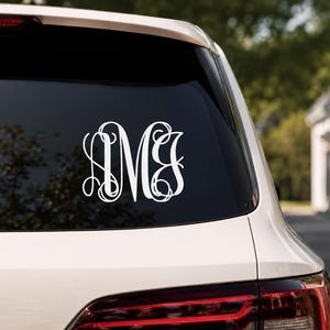 May include: A white vinyl decal featuring a decorative three-letter monogram with the letters L, M, and B in a stylized, interlocking script font. The decal is applied to the center of a dark tinted rear vehicle window on a white car.