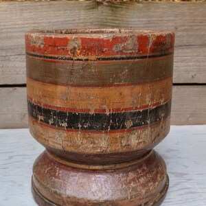 May include: A wooden mortar and pestle with a wide base and a cylindrical top. The mortar is painted with a pattern of red, black, and green stripes. The mortar is weathered and shows signs of age.
