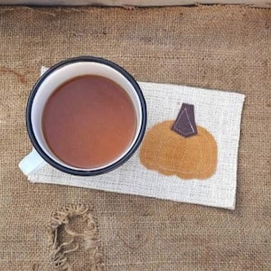 May include: A white enamel mug filled with brown liquid sits on a rectangular coaster featuring an orange pumpkin graphic with a brown stem. The coaster rests on a piece of brown burlap inside a weathered, rectangular wooden tray with side handles.