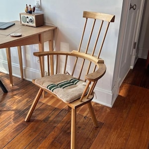 Op de afbeelding: Een houten Windsor-stoel met een gebogen rug en armleuningen. De stoel heeft een lichte houten afwerking en een zitkussen met groene strepen. De stoel staat in een kamer met houten vloeren en een houten bureau.