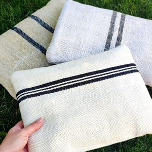 Pode incluir: Três almofadas retangulares com um padrão de tecido em espinha. Uma almofada é branco-sujo com listras pretas, outra é cinza claro com listras cinza escuro, e a terceira é bege com listras azul marinho. As almofadas estão na grama verde.