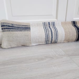 May include: A rectangular doorstop made of fabric with a patchwork design. The doorstop features sections of beige, white, and beige fabric with navy blue stripes. It rests on a light gray wooden floor.