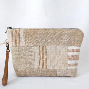 Puede incluir: Bolso de mano beige con patchwork, cremallera negra y correa de muñeca de cuero marrón. El bolso rectangular presenta varios patrones de tejido, incluyendo rayas verticales y diseños de espiga. El bolso mide unos 25 cm de ancho.