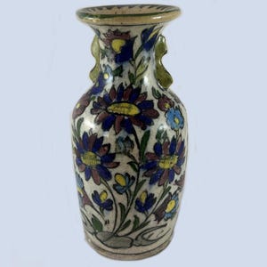 May include: A small, hand-painted ceramic vase with a cream-colored base. The vase is adorned with blue, purple, and yellow floral patterns. It has two small handles and a green rim. The vase is approximately 6 inches tall.