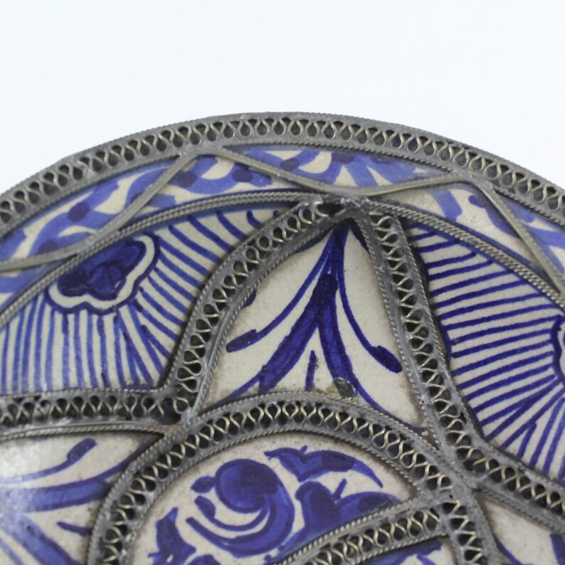 K&ouml;nnte beinhalten: Ein wei&szlig;er Keramikteller mit einem blauen und wei&szlig;en Blumenmuster. Der Teller ist mit einem silbernen Metallrand verziert.