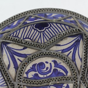 K&ouml;nnte beinhalten: Ein wei&szlig;er Keramikteller mit einem blauen und wei&szlig;en Blumendesign. Der Teller ist mit einem silbernen Metallrand verziert.