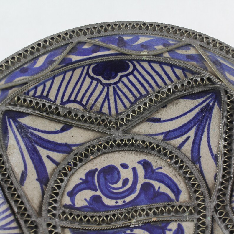K&ouml;nnte beinhalten: Ein wei&szlig;er Keramikteller mit einem blauen und wei&szlig;en Blumendesign. Der Teller ist mit einem silbernen Metallrand verziert.