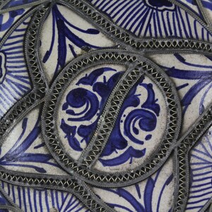 K&ouml;nnte beinhalten: Nahaufnahme einer Keramikfliese mit einem blauen und wei&szlig;en Blumenmuster. Die Fliese ist mit einem silbernen Metallrand verziert.