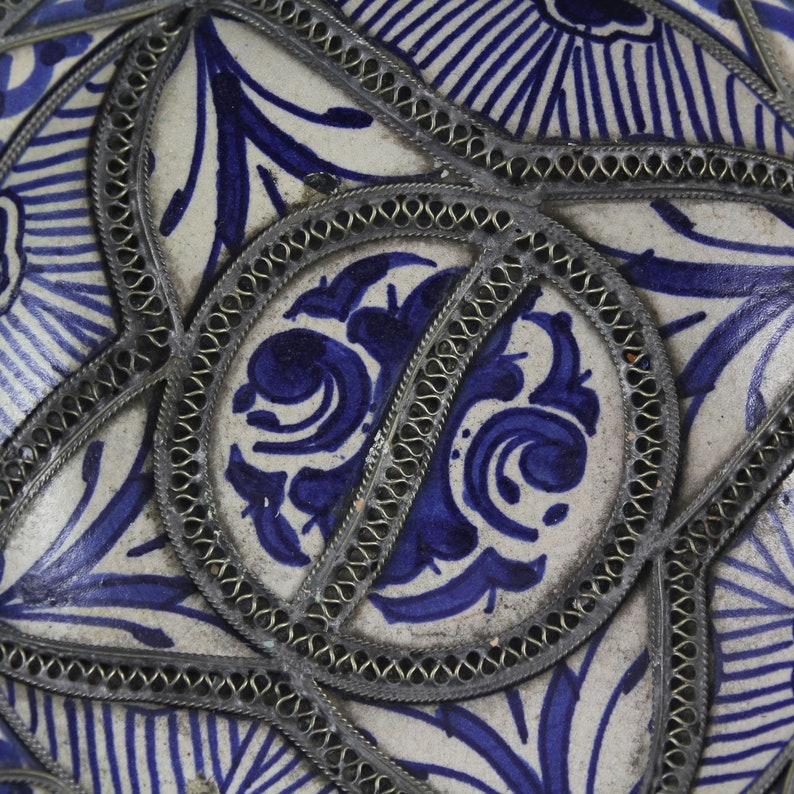 K&ouml;nnte beinhalten: Nahaufnahme einer Keramikfliese mit einem blauen und wei&szlig;en Blumenmuster. Die Fliese ist mit einem silbernen Metallrand verziert.