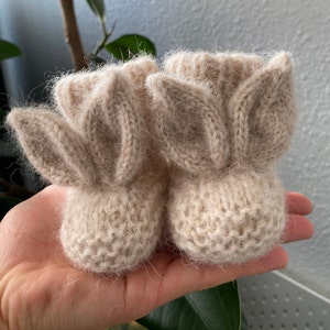 Puede incluir: Un par de patucos de punto en color beige con estructuras puntiagudas en forma de orejas en la parte delantera. Los patucos están confeccionados con un hilo suave y difuso, con un puño acanalado en la parte superior y una puntera redondeada, mostrando una construcción de punto hecho a mano.