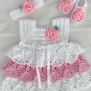 May include: A white crocheted baby dress with pink and white ruffles, a matching headband with a pink flower, and a pair of pink crocheted baby shoes with flowers.