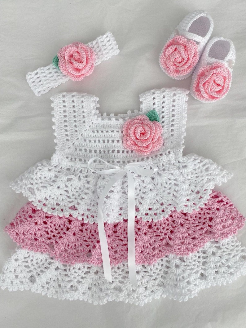 May include: A white crocheted baby dress with pink and white ruffles, a matching headband with a pink flower, and a pair of pink crocheted baby shoes with flowers.
