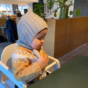 May include: A young child wearing a light blue and white knitted hat with a pointed top sits in a high chair at a table. The child is wearing a brown and white patterned sweater. The table has a green surface.