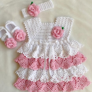 May include: A white crocheted baby dress with pink ruffles and a white satin ribbon. The dress has a pink flower on the bodice and comes with a matching headband and shoes.