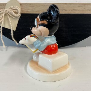 Puede incluir: Una figura de cer&aacute;mica de Mickey Mouse, con cabeza negra, gafas con montura marr&oacute;n y atuendo rojo y blanco. La figura est&aacute; leyendo un libro y est&aacute; montada sobre una base blanca. El fondo incluye un coraz&oacute;n de madera con un lazo de cuerda.