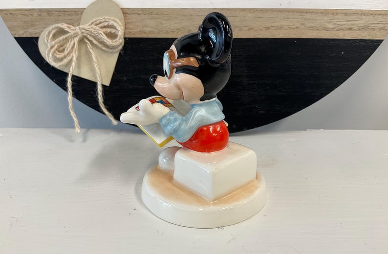 Puede incluir: Una figura de cer&aacute;mica de Mickey Mouse, con cabeza negra, gafas con montura marr&oacute;n y atuendo rojo y blanco. La figura est&aacute; leyendo un libro y est&aacute; montada sobre una base blanca. El fondo incluye un coraz&oacute;n de madera con un lazo de cuerda.