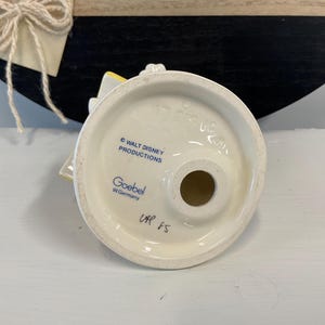 Puede incluir: Base de figura de cer&aacute;mica blanca con dise&ntilde;o circular, con el texto "WALT DISNEY PRODUCTIONS" y "Goebel W. Germany". La base tiene un agujero en el centro y el texto manuscrito "UAP 85".