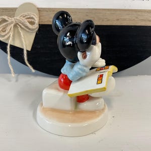 Puede incluir: Una figura de cer&aacute;mica de Mickey Mouse, con pantalones cortos rojos, guantes blancos y una camisa azul claro. Mickey sostiene un bloc de notas blanco con cuadrados de colores. La figura est&aacute; sobre una base blanca.