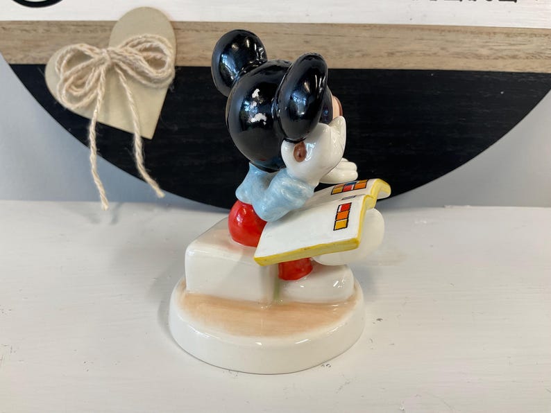 Puede incluir: Una figura de cer&aacute;mica de Mickey Mouse, con pantalones cortos rojos, guantes blancos y una camisa azul claro. Mickey sostiene un bloc de notas blanco con cuadrados de colores. La figura est&aacute; sobre una base blanca.
