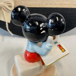 Puede incluir: Una figura vintage de Mickey Mouse. La figura con orejas negras lleva una camisa azul claro y pantalones cortos rojos, sosteniendo un bloc de notas blanco con cuadrados de colores. La figura est&aacute; sobre una base blanca.