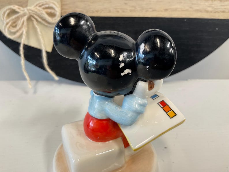 Puede incluir: Una figura vintage de Mickey Mouse. La figura con orejas negras lleva una camisa azul claro y pantalones cortos rojos, sosteniendo un bloc de notas blanco con cuadrados de colores. La figura est&aacute; sobre una base blanca.