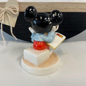 Puede incluir: Una figura de cer&aacute;mica de Mickey Mouse, con orejas negras, torso rojo y cuello azul. La figura sostiene un libro blanco con cuadrados coloridos. Est&aacute; sobre una base blanca con borde beige.
