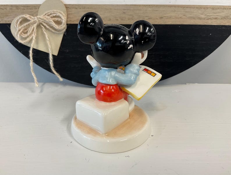 Puede incluir: Una figura de cer&aacute;mica de Mickey Mouse, con orejas negras, torso rojo y cuello azul. La figura sostiene un libro blanco con cuadrados coloridos. Est&aacute; sobre una base blanca con borde beige.