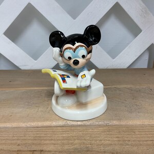 Puede incluir: Una figura de cer&aacute;mica de Mickey Mouse, con las ic&oacute;nicas orejas negras, gafas y una camisa azul. Mickey posa pensativamente, apoyado en una base blanca con un libro colorido. La figura est&aacute; sobre una superficie de madera.
