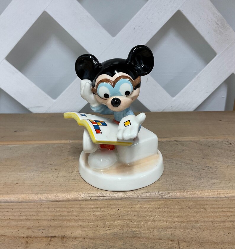 Puede incluir: Una figura de cer&aacute;mica de Mickey Mouse, con las ic&oacute;nicas orejas negras, gafas y una camisa azul. Mickey posa pensativamente, apoyado en una base blanca con un libro colorido. La figura est&aacute; sobre una superficie de madera.
