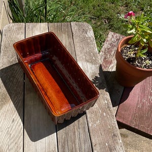 May include: A rectangular ceramic planter with a glossy, deep brown glaze and a vertical ribbed texture on the exterior walls. The interior is smooth and finished in a matching dark brown tone, featuring a deep basin for holding soil or plants.