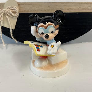 Puede incluir: Una figura de cer&aacute;mica de Mickey Mouse con gafas y una camisa azul claro, sosteniendo un libro con formas geom&eacute;tricas coloridas. La figura est&aacute; sobre una base blanca con una superficie de color beige claro.
