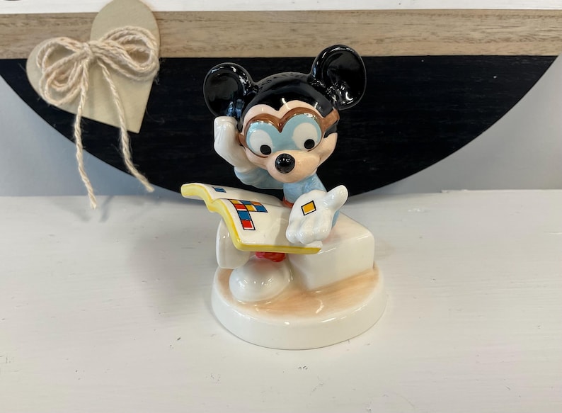 Puede incluir: Una figura de cer&aacute;mica de Mickey Mouse con gafas y una camisa azul claro, sosteniendo un libro con formas geom&eacute;tricas coloridas. La figura est&aacute; sobre una base blanca con una superficie de color beige claro.
