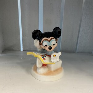 Puede incluir: Una figura de cer&aacute;mica de Mickey Mouse, con orejas negras, una camisa azul y guantes blancos. Mickey sostiene un libro y parece estar pensando. La figura est&aacute; sobre una base blanca con un acento marr&oacute;n claro.