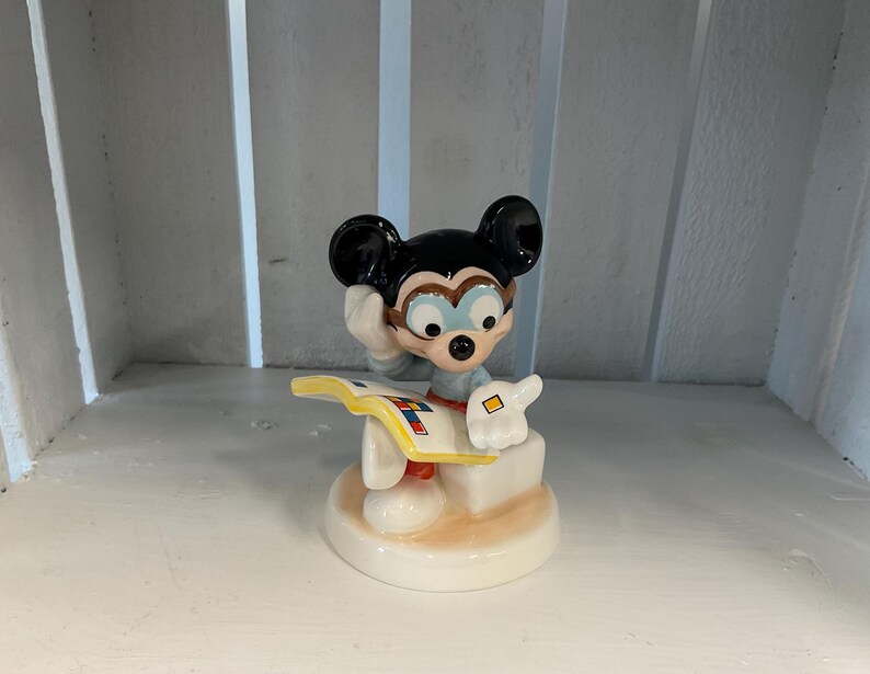 Puede incluir: Una figura de cer&aacute;mica de Mickey Mouse, con orejas negras, una camisa azul y guantes blancos. Mickey sostiene un libro y parece estar pensando. La figura est&aacute; sobre una base blanca con un acento marr&oacute;n claro.