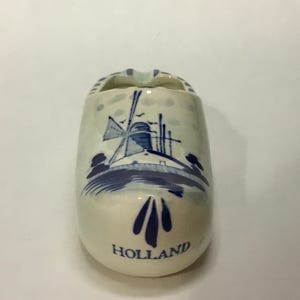 May include: A ceramic Dutch clog ashtray, painted white with a blue windmill scene and the word "HOLLAND". The ashtray has a small opening at the top and is a decorative souvenir.