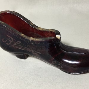 May include: A dark red glass shoe with a high heel. The shoe has a gold inscription that reads "J. Blake 1910".
