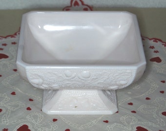 May include: A square, footed, pink milk glass bowl with a raised floral design. The bowl is empty and sits on a white lace doily with red hearts.
