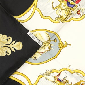 May include: A black and white silk scarf with a gold trim. The scarf features a white horse with a gold bridle and a blue and red rider. The scarf also features a gold floral design. The scarf has a label that reads 'Fabrique en France Made in France 100% Silk Dry Clean Only'. 