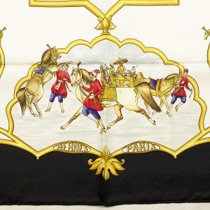 May include: A white silk scarf with a black border and a gold design. The center of the scarf features a scene of people riding horses in a procession. The people are wearing colorful clothing and the horses are white with gold accents. The text 'HERMES PARIS' is written in gold letters at the bottom of the scarf.