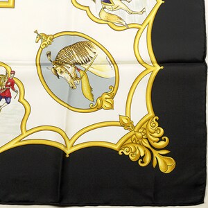 May include: A white and black silk scarf with a gold and yellow design. The design features a horse, a rider, and a floral motif. The scarf is folded in half and the design is visible on both sides.