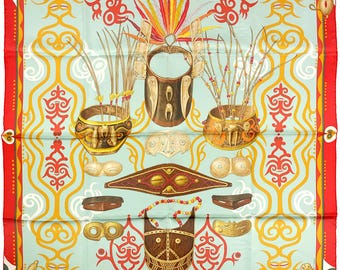 May include: Square silk scarf with a light blue background and a red border. The scarf features a repeating pattern of tribal masks, decorative bowls, and feather headdresses in shades of brown, yellow, and red. The scarf has the text "HERMES PARIS" at the bottom.