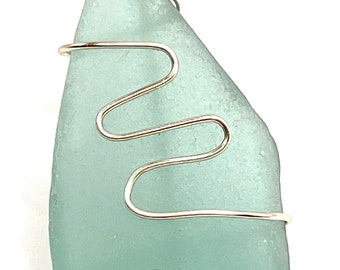 May include: A light blue sea glass pendant wrapped in silver wire. The wire is twisted and forms a wavy pattern around the glass.