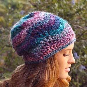 May include: A multi-coloured, hand-crocheted beanie in shades of teal, purple, and pink. The hat is worn by a person with long, wavy auburn hair and a blue shirt. The beanie is a stylish accessory for cooler weather.