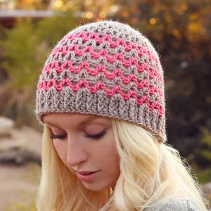 May include: A handmade crochet beanie in a neutral beige and pink color scheme. The hat features a textured pattern and is designed to fit snugly. The beanie is a stylish accessory for cooler weather, offering both warmth and a unique aesthetic.