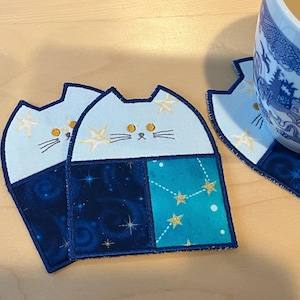 Puede incluir: Tres posavasos con forma de gato azul y blanco con un diseño de noche estrellada. Los posavasos están hechos de tela y tienen una textura acolchada.